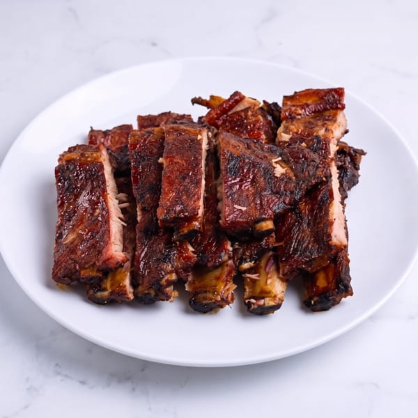 Glazed, charred barbecue ribs stacked on a white plate atop a marble surface.