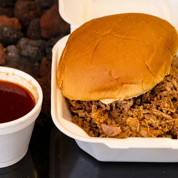 Pulled pork sandwich in a white takeout container with a side of barbecue sauce.