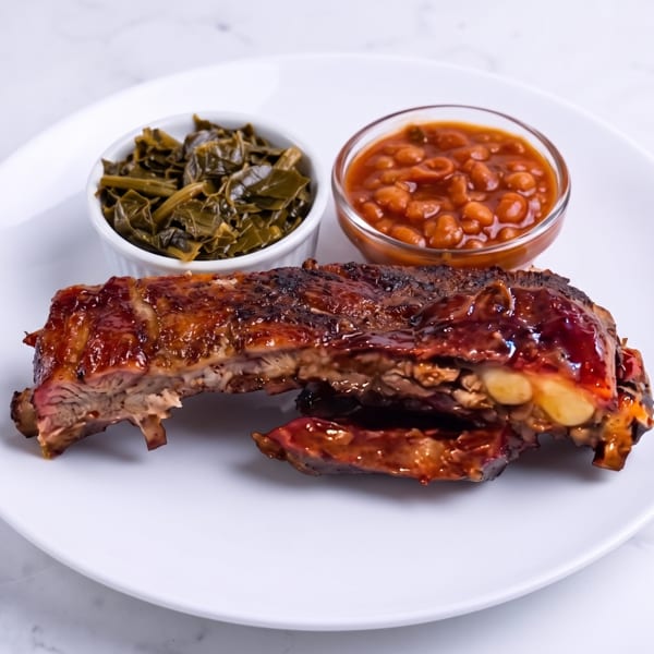 Glazed barbecue ribs served with collard greens and baked beans on a white plate.
