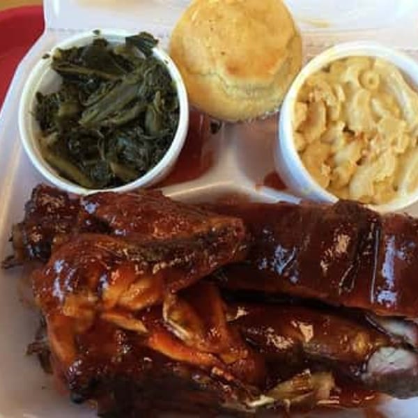 BBQ chicken and ribs with collard greens, macaroni and cheese, and a roll in a takeout container.