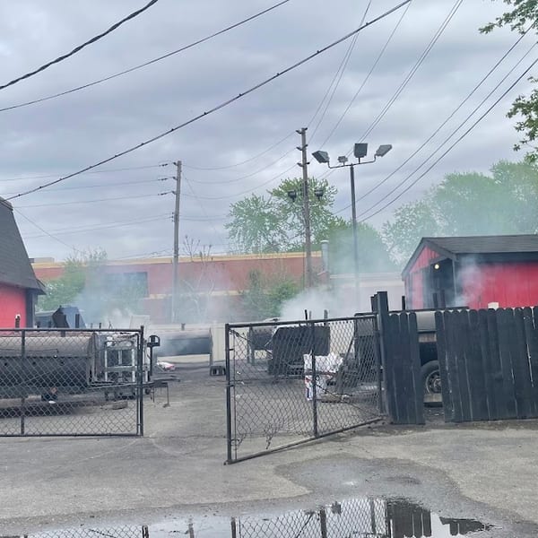 Smoke billows from multiple large barbecue smokers behind a red building with black roofs.